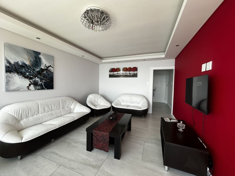 A well-maintained living room featuring large format floor tiles, a bold red feature wall, and recessed ceiling lighting.