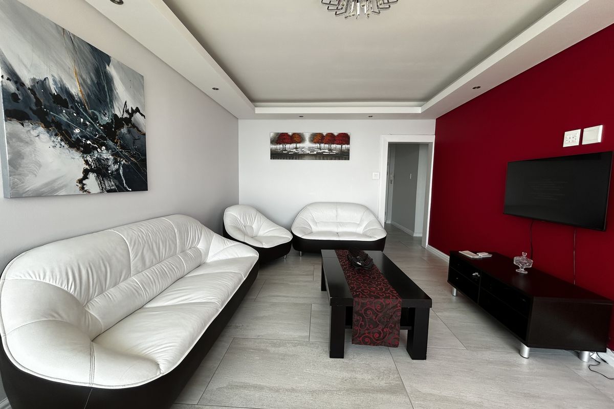A well-maintained living room featuring a modern recessed ceiling with downlights, neutral tiled flooring, and a bold red accent wall.
