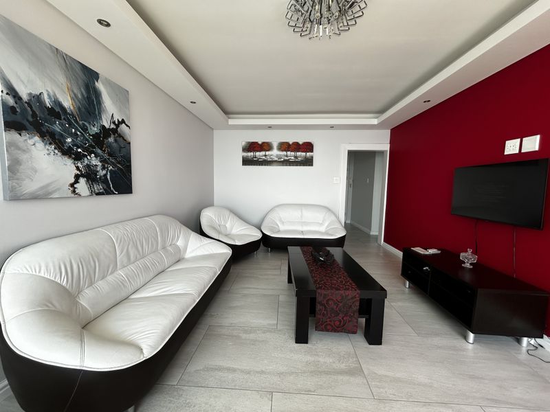 A well-maintained living room featuring a modern recessed ceiling with downlights, neutral tiled flooring, and a bold red accent wall.