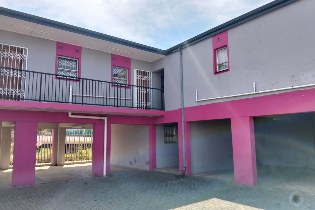 Exterior view of an apartment complex featuring a paved parking area, covered bays, and a balcony with security gates and railings.