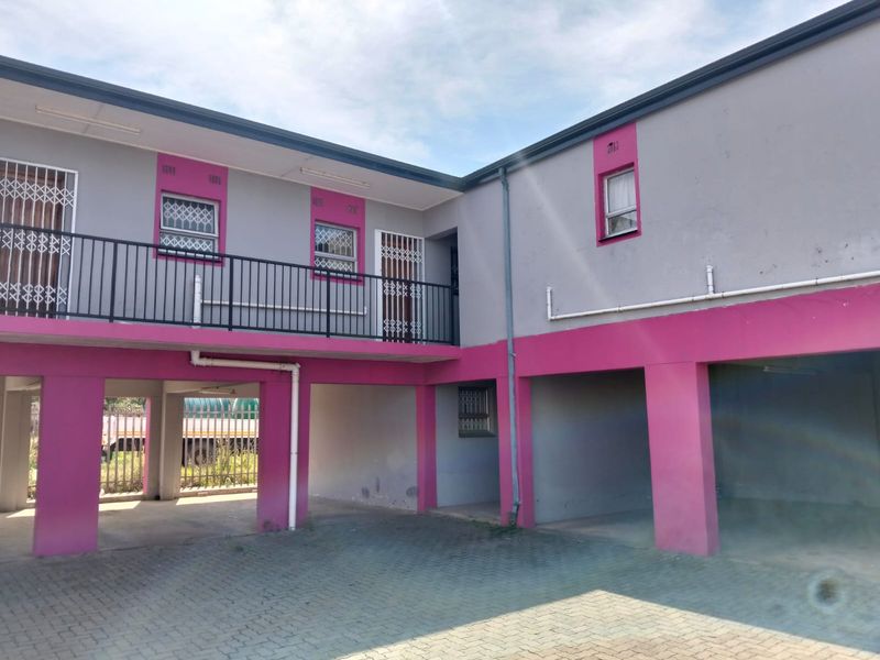 Exterior view of an apartment complex featuring a paved parking area, covered bays, and a balcony with security gates and railings.
