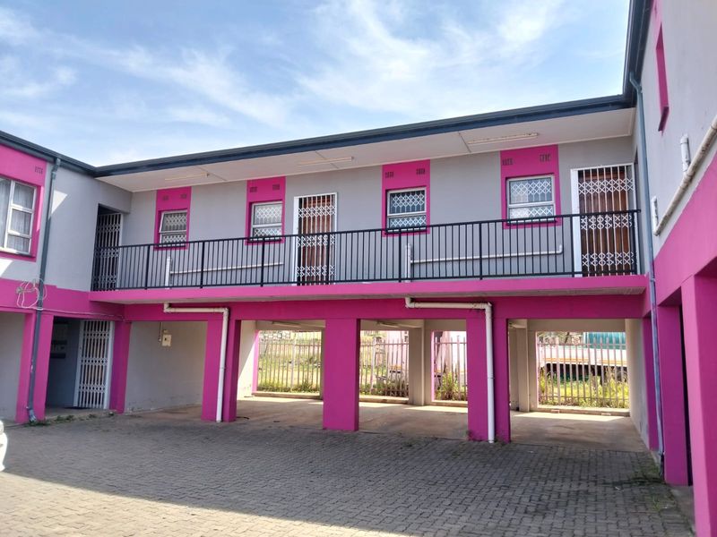 Exterior view of an apartment building featuring a paved driveway, covered parking bays, and an upper-level balcony with security gates.