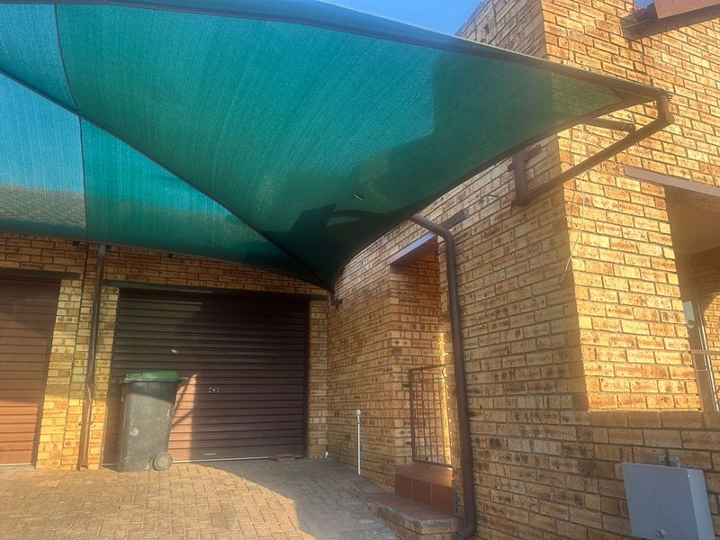 Front exterior view of a townhouse featuring a brick facade, a roller garage door, and a green shade sail covering the driveway area.