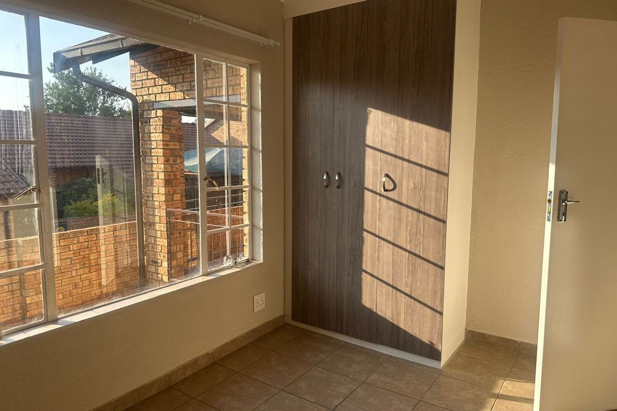 A bright, empty bedroom featuring tiled flooring, a built-in wooden wardrobe, and large windows with security bars.