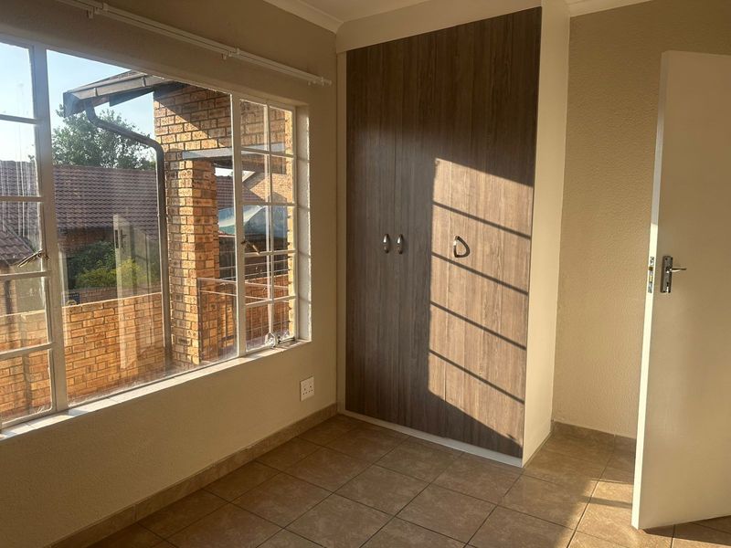 A bright, empty bedroom featuring tiled flooring, a built-in wooden wardrobe, and large windows with security bars.
