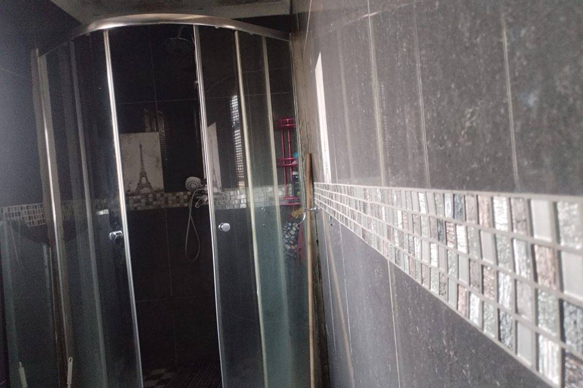 A bathroom featuring a curved glass shower enclosure and dark wall tiles with a decorative mosaic border, showing signs of ceiling water damage.
