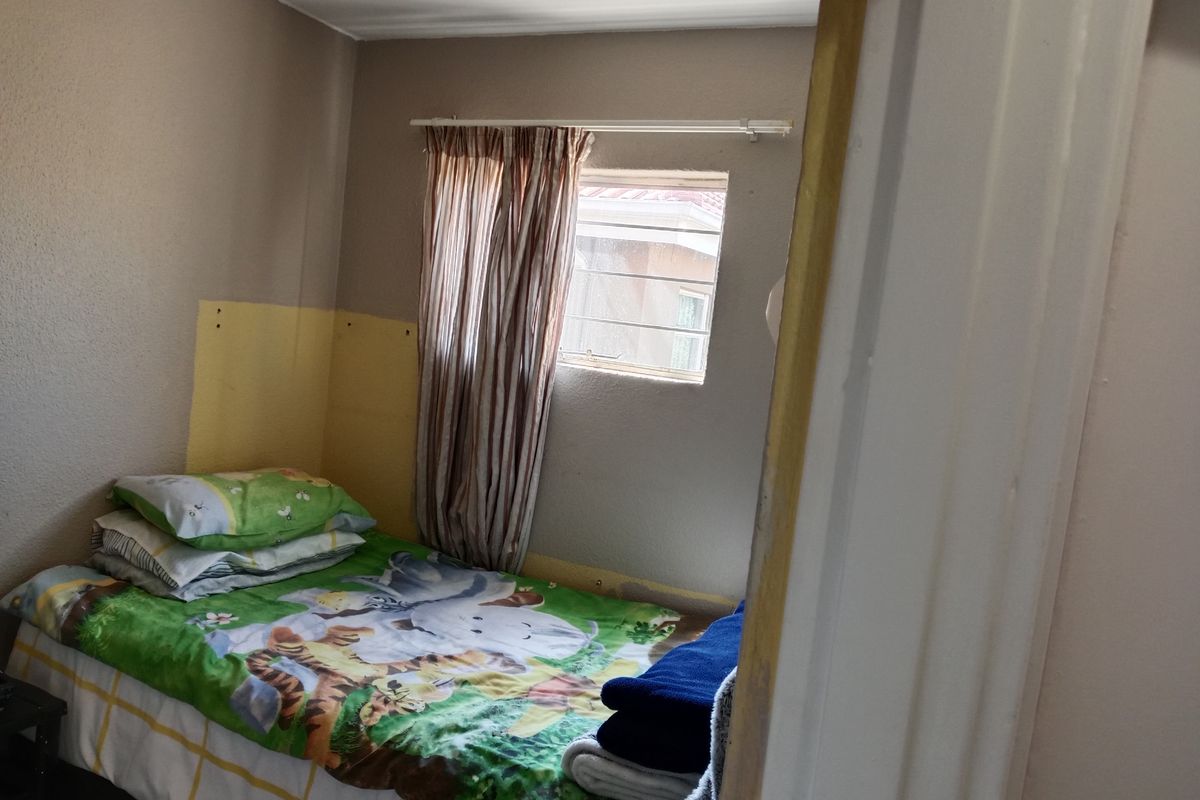 A compact bedroom featuring a window with security bars and visible wall surface wear, suitable for renovation or cosmetic updates.