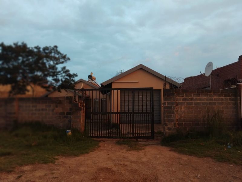 Front exterior view of a single-story house featuring a metal security gate, perimeter wall with razor wire, and a solar water heater installation.
