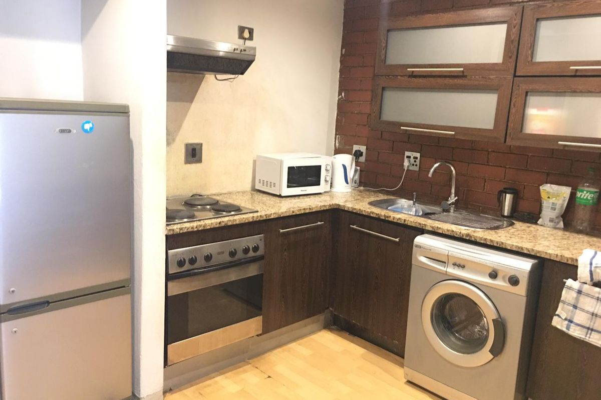 Compact kitchen featuring granite countertops, built-in oven, electric hob, extractor fan, and dark wood cabinetry with an exposed brick feature wall.