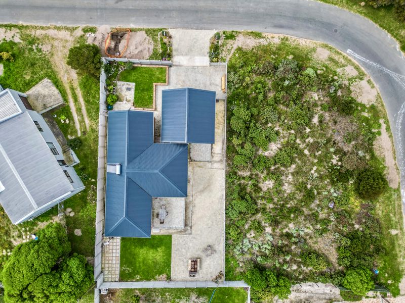 Aerial view of the property showing the house layout, metal roof, paved driveway, and surrounding garden area.