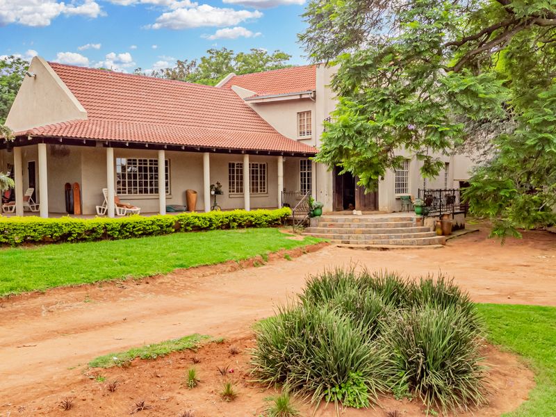 A spacious rear exterior view of the small holding featuring a large covered patio, tiled roof, and stone entrance steps set within a landscaped garden.