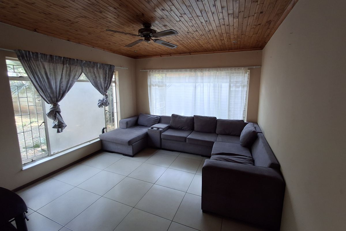 A living room featuring tiled flooring, a timber tongue and groove ceiling with a ceiling fan, and large windows with security bars.