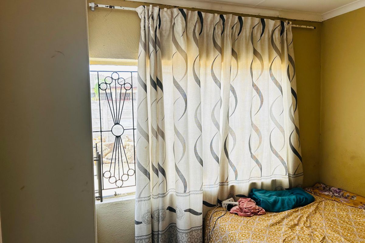 A bedroom featuring a window with a decorative security gate and a curtain rod, showing signs of wear on the wall and fixtures.