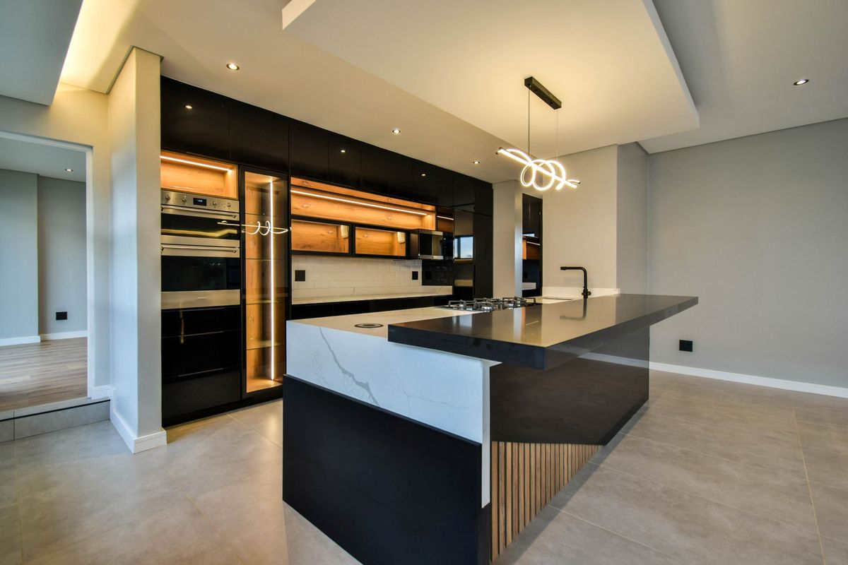 Modern kitchen featuring high-gloss black cabinetry, a large stone-topped island with integrated hob, built-in ovens, and stylish recessed and pendant lighting.
