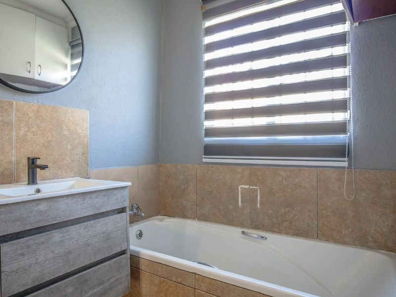 A functional bathroom featuring a modern vanity with basin, a bathtub with tiled surround, and a roller blind for privacy.