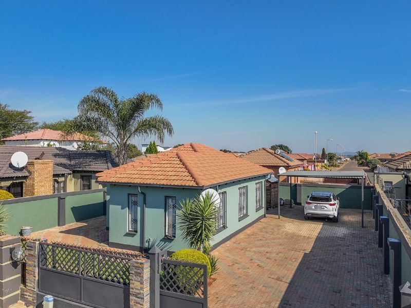 An elevated view of a well-maintained residential property featuring a tiled roof, paved driveway, secure perimeter walling, and a covered carport.