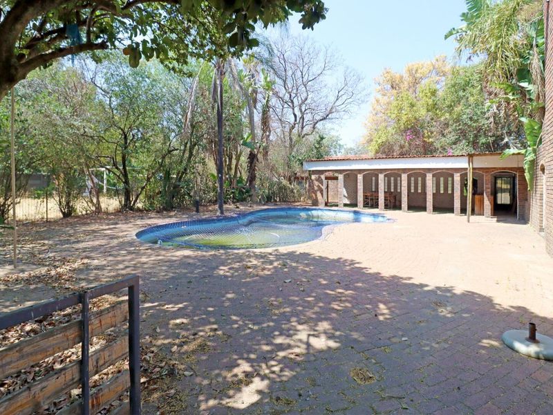 Outdoor pool area featuring a paved patio, covered entertainment space with brick columns, and mature garden surroundings.