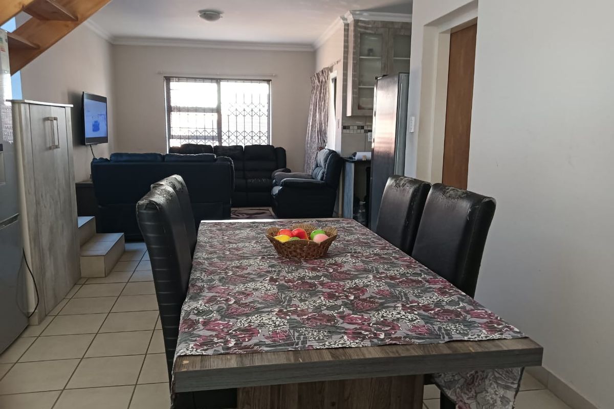 Open-plan living and dining area featuring tiled floors, a wooden staircase, and integrated kitchen cabinetry with security bars on the window.