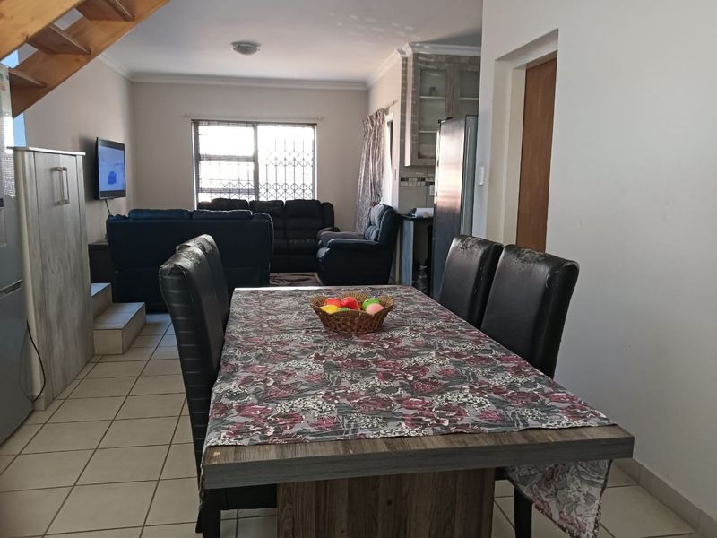 Open-plan living and dining area featuring tiled floors, a wooden staircase, and integrated kitchen cabinetry with security bars on the window.