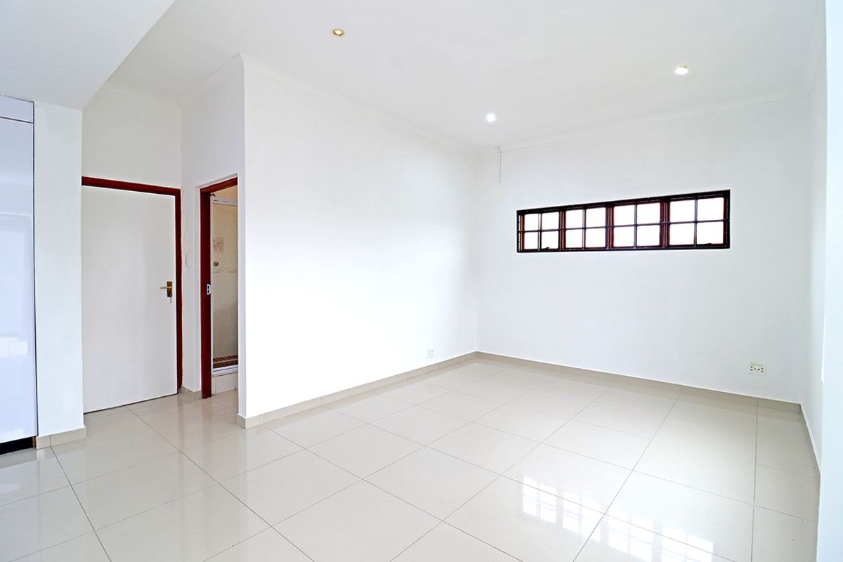 A bright, empty bedroom featuring neutral tiled flooring, recessed ceiling lighting, and a horizontal window with dark frames.