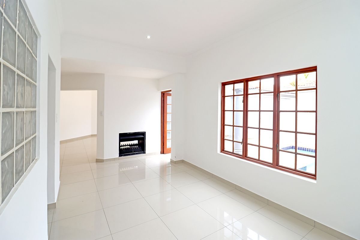 A bright, empty living room featuring large tiled flooring, a built-in fireplace, and classic wooden-framed windows overlooking the exterior.