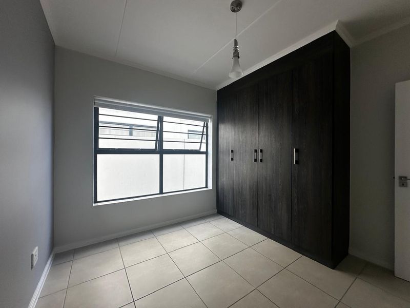 A clean, empty bedroom featuring neutral tiled flooring, a large window with security bars, and dark wood built-in cupboards.
