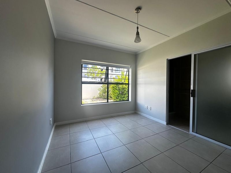 A bright, empty bedroom featuring neutral tiled flooring, a large window with security bars, and a sliding door leading to an adjacent space.