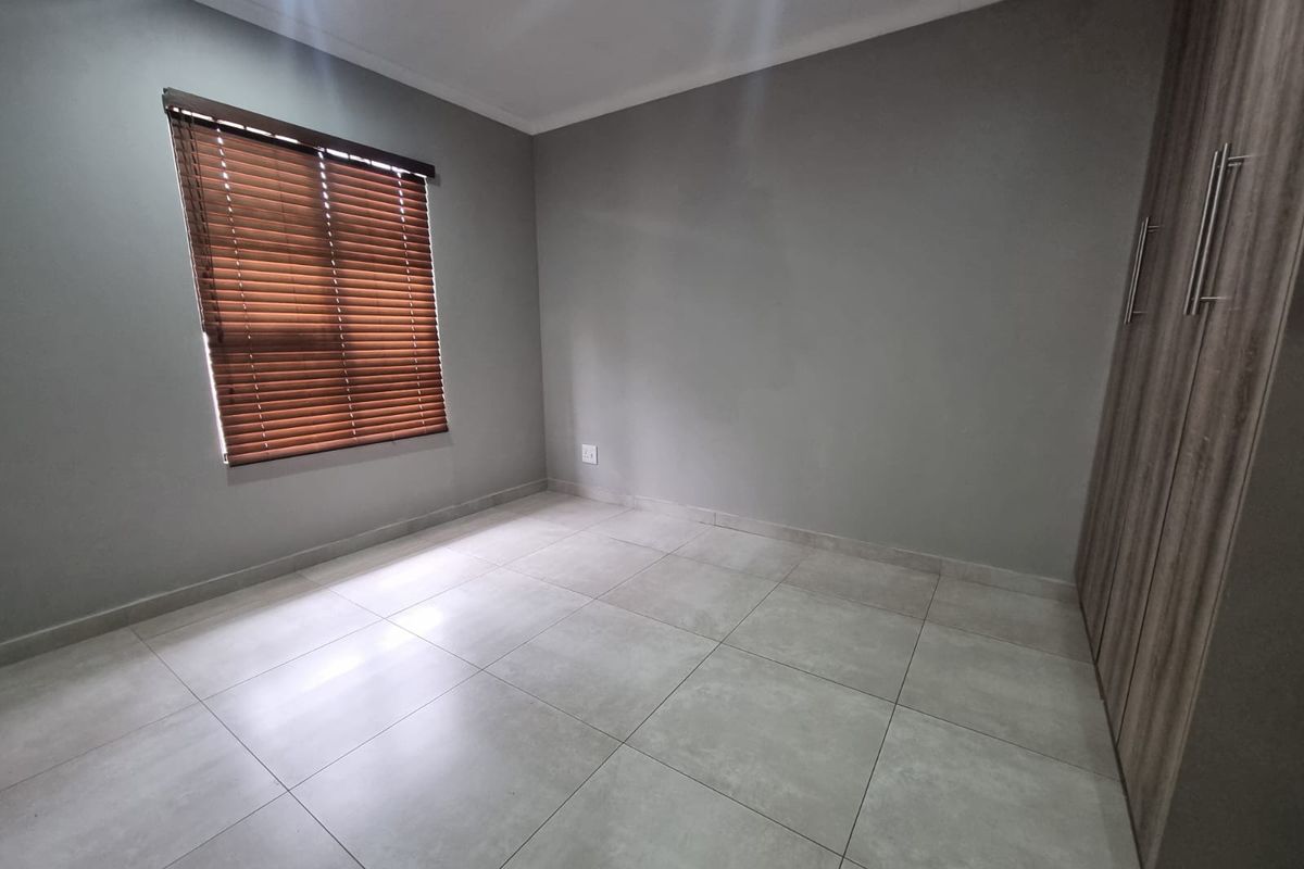 A clean, empty bedroom featuring neutral grey walls, large format floor tiles, and built-in wooden cabinetry with modern handles.