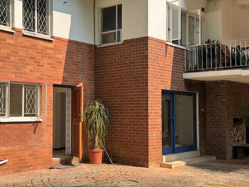 Rear exterior view of a brick cottage featuring a paved courtyard, balcony with metal railings, and multiple access points.