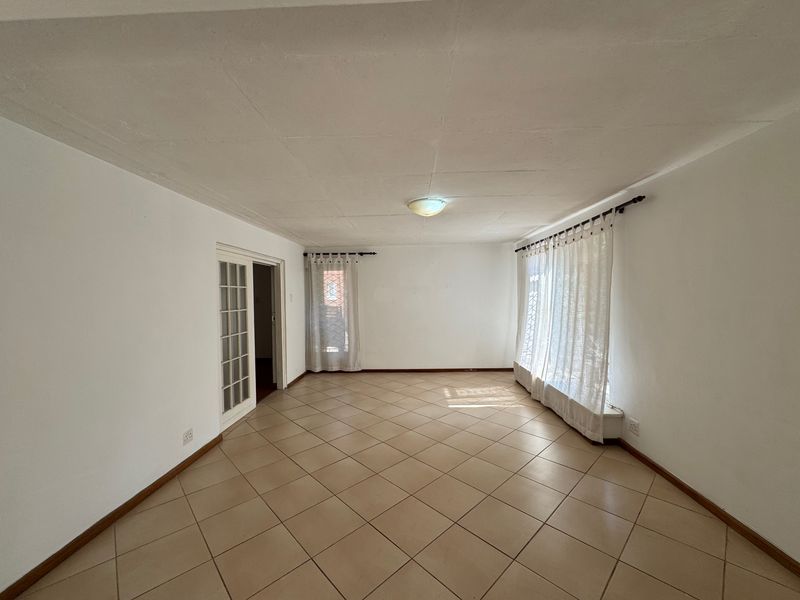 A spacious, empty living room featuring neutral tiled flooring, a classic french door, and natural light through large windows.