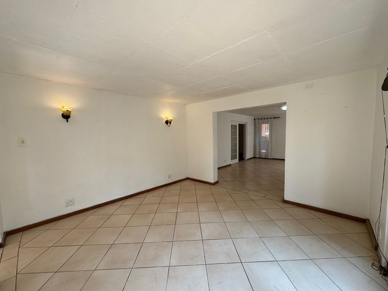 An empty, open-plan living area featuring neutral tiled flooring, wall-mounted lighting, and direct access to an adjoining room through a wide opening.