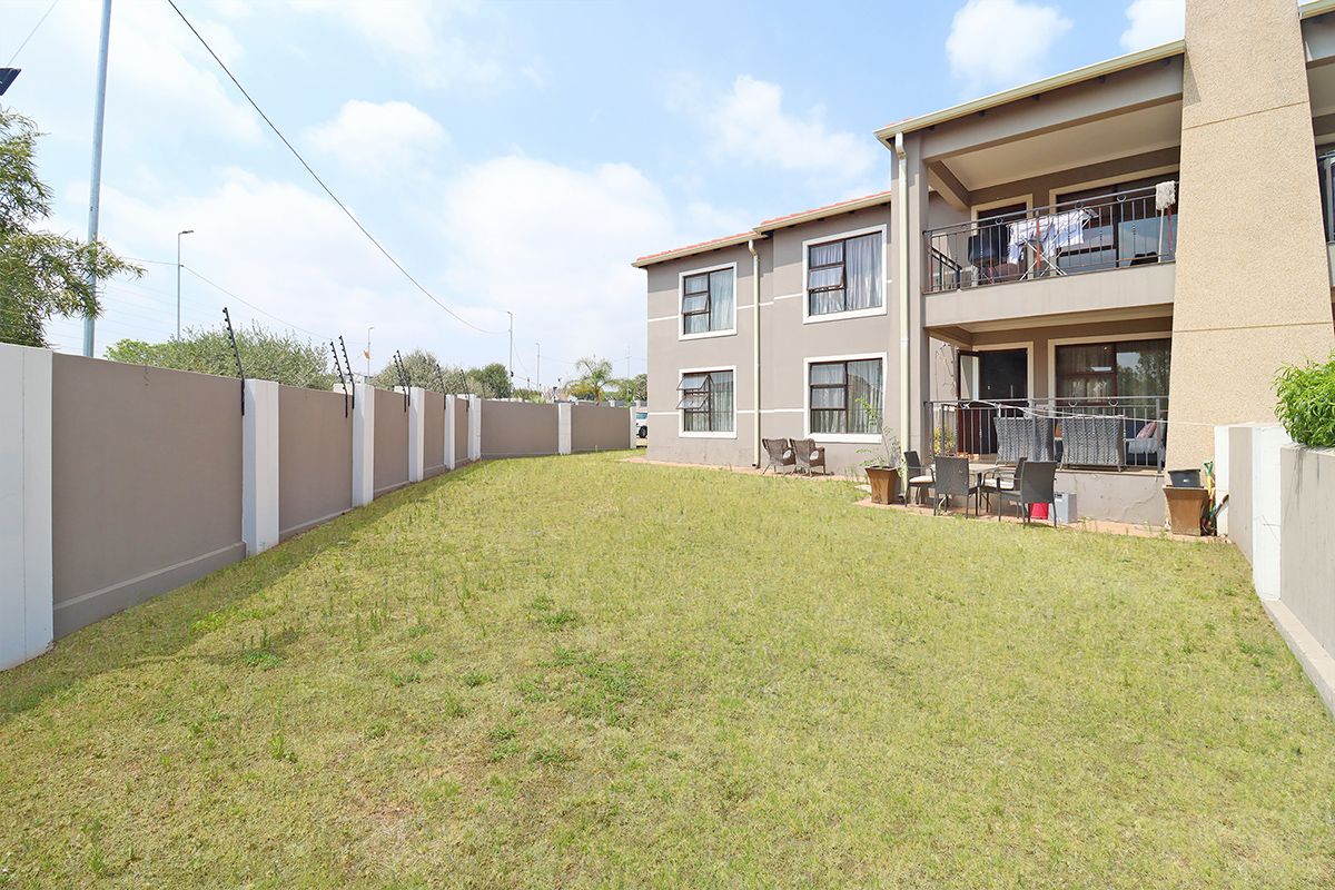 A view of the rear garden and patio area of the apartment complex, featuring a lawn, perimeter wall with electric fencing, and balcony access.