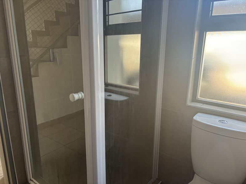 A functional bathroom featuring a toilet, shower enclosure with glass door, and tiled finishes, illuminated by natural light through a frosted window.