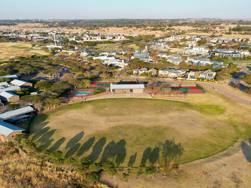 Aerial view of a residential estate featuring a large sports field, tennis courts, swimming pool, and clubhouse facilities within a landscaped community setting.