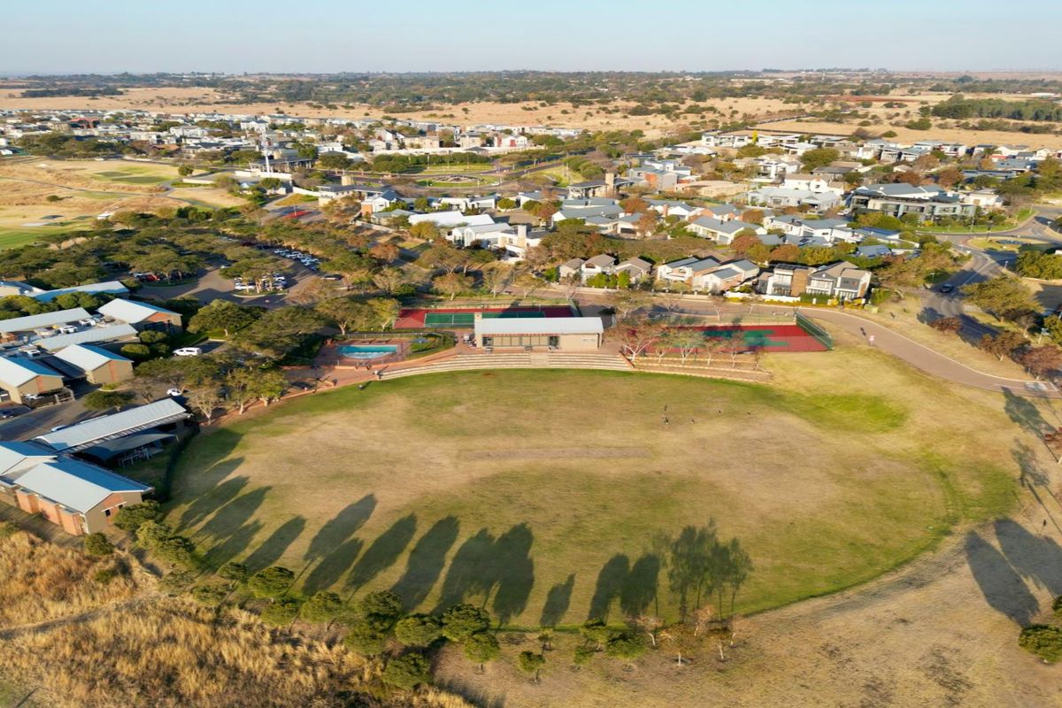 Aerial view of a residential estate featuring a large sports field, tennis courts, swimming pool, and clubhouse facilities within a landscaped community setting.