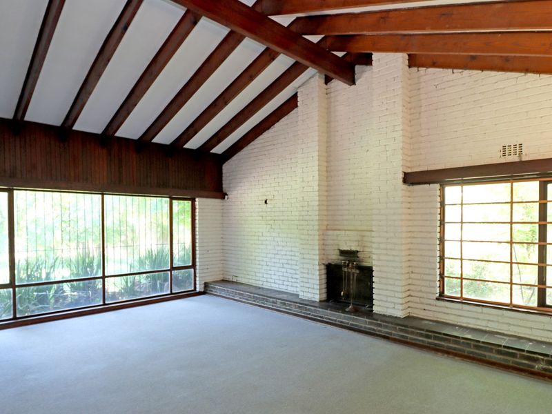 Spacious living room featuring exposed timber ceiling beams, a painted brick fireplace, and large windows providing ample natural light.