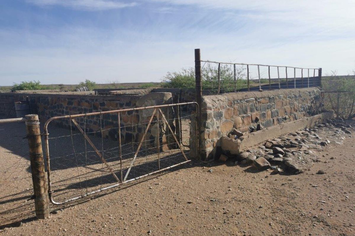An outdoor farm enclosure featuring rustic stone walls, a metal gate, and a concrete trough, showing signs of age and weathering.