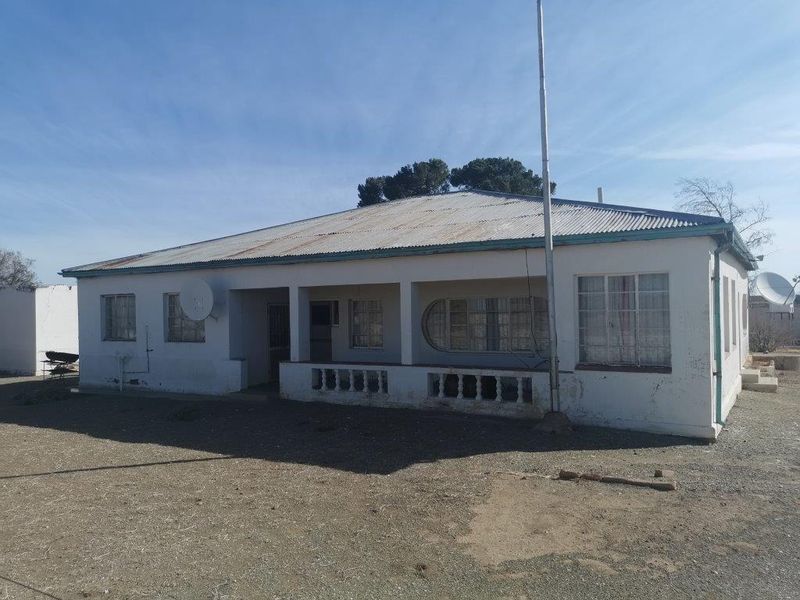 Front elevation of a farm-style dwelling featuring a covered veranda, corrugated metal roof, and concrete balustrade, set on a large open plot.
