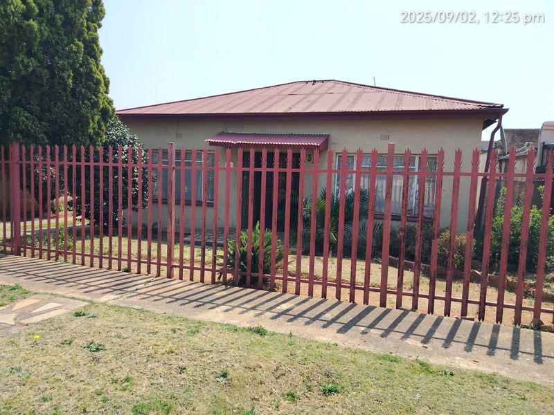 Front elevation of a single-story house featuring a corrugated metal roof, entrance awning, and perimeter metal security fencing.
