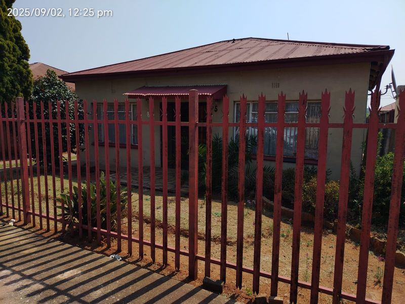 Front view of a single-story house featuring a corrugated metal roof, security fencing, and window bars, set behind a metal palisade perimeter fence.