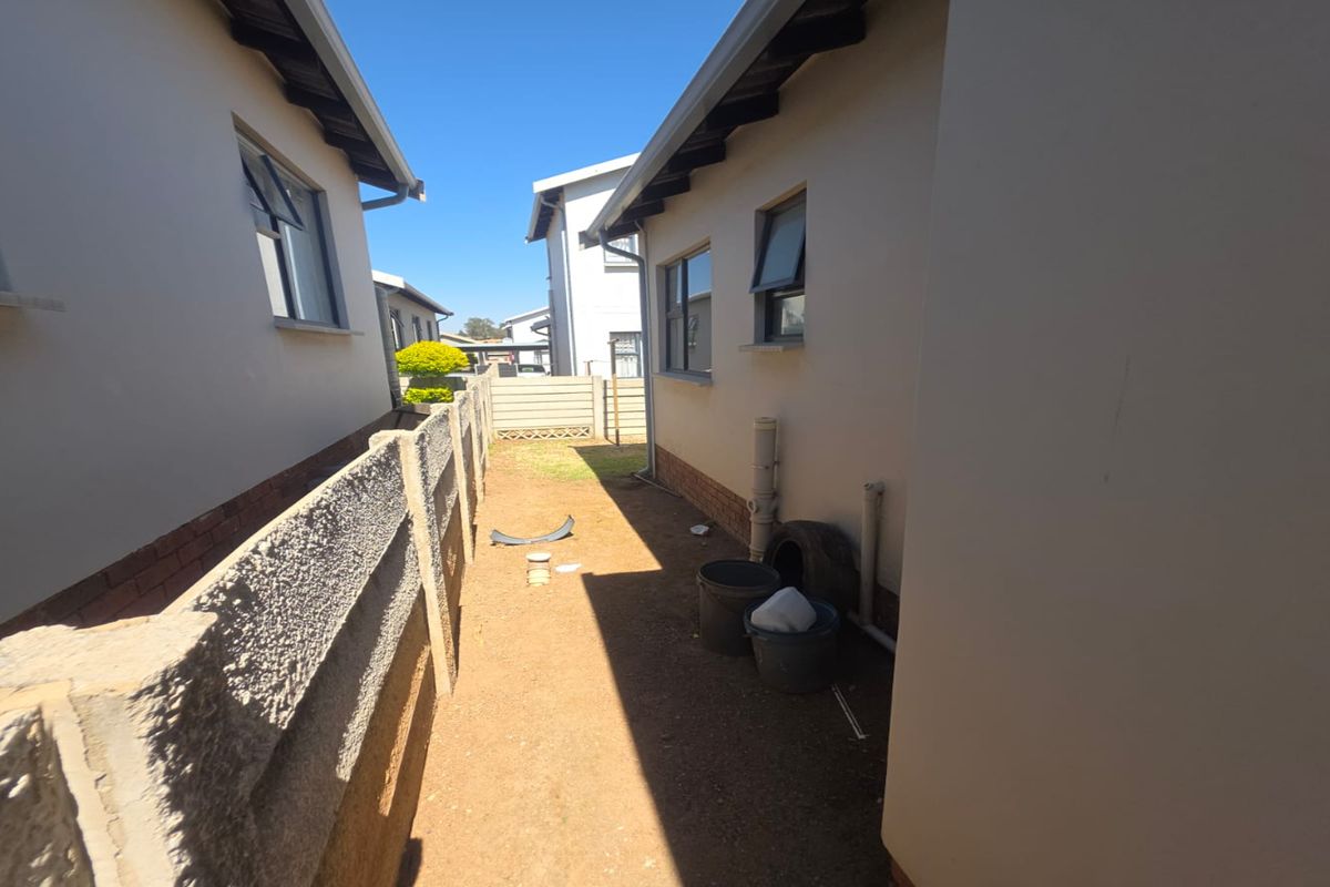 A narrow side exterior passage between two townhouse units featuring exposed plumbing and an unpaved ground surface.
