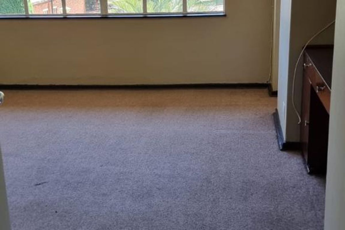 An empty bedroom featuring a large metal-framed window, fitted carpeting, and a built-in desk area, offering a blank canvas for renovation.