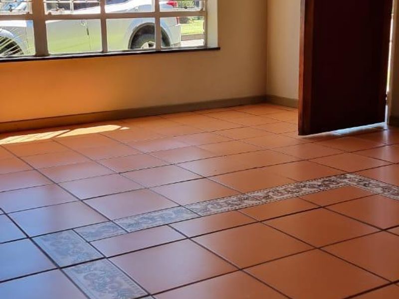 An empty living room featuring terracotta-style tiled flooring with decorative border inlay and large windows fitted with security bars.