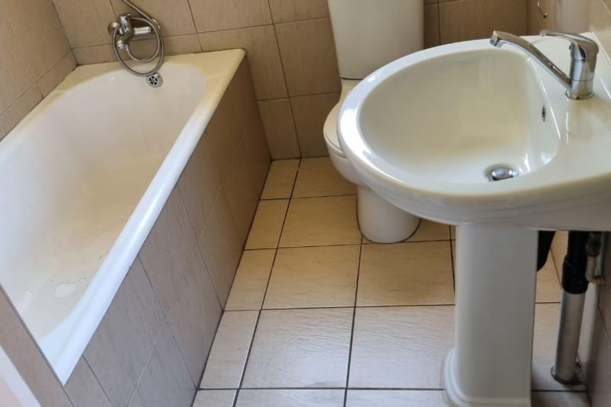 A compact bathroom featuring a pedestal basin, toilet, and bathtub with full-height wall tiling and floor tiles, requiring some cosmetic updates.