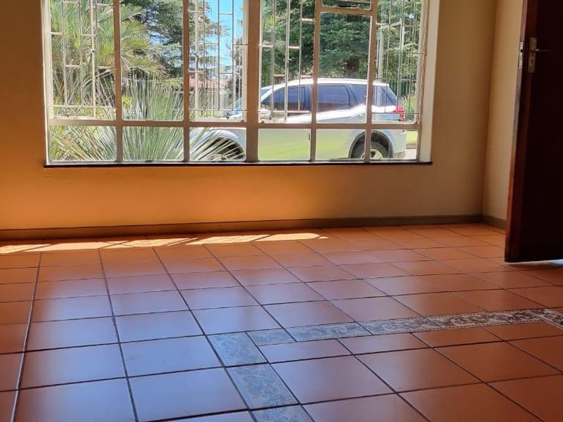 An empty living room featuring tiled flooring with a decorative border and large windows fitted with security bars.