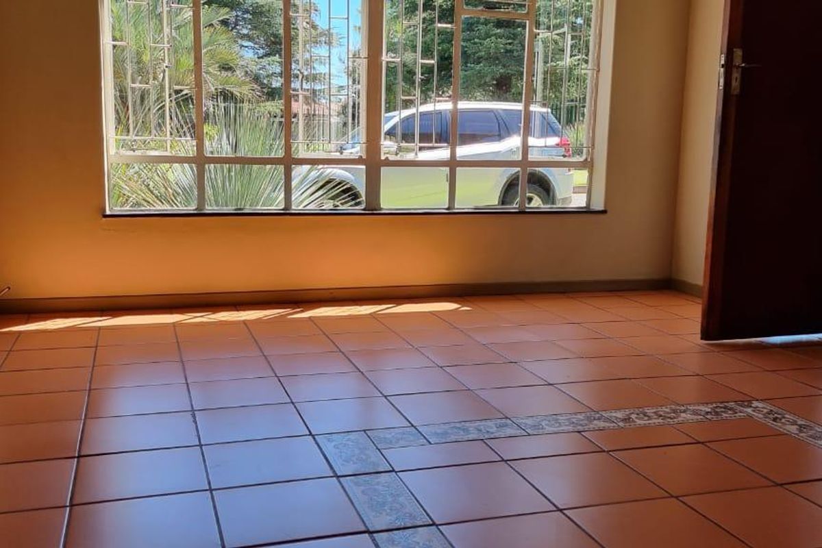 An empty living room featuring tiled flooring with a decorative border and large windows fitted with security bars.