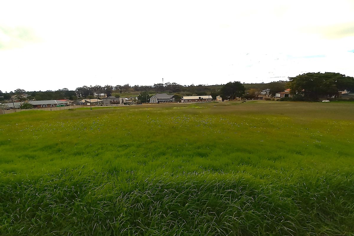 A wide-angle view of a vacant residential plot featuring open grassland with surrounding neighborhood buildings in the distance.