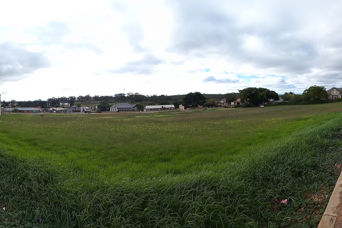 A wide-angle view of a vacant residential corner plot featuring established grass, adjacent paved sidewalks, and street-side infrastructure.