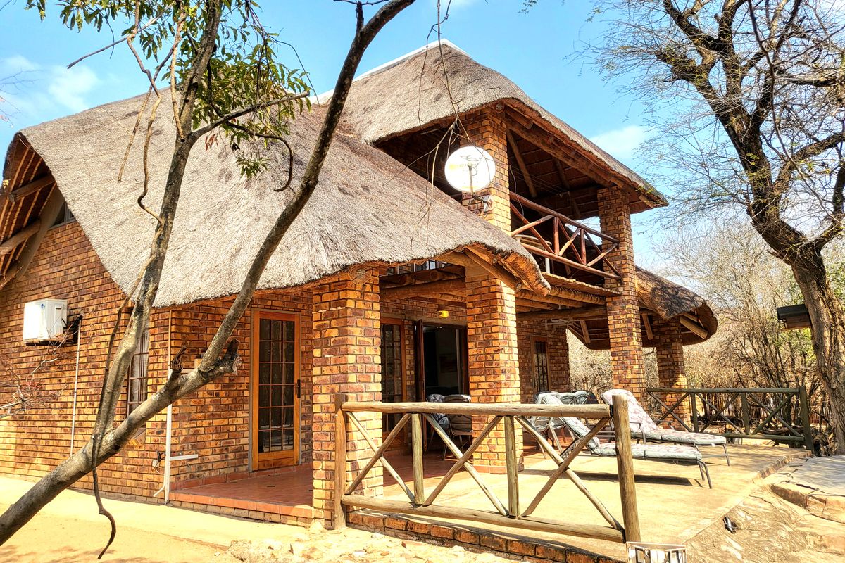 A rustic double-story house featuring a traditional thatched roof, exposed brick exterior, and a spacious covered patio with a wooden balcony above.