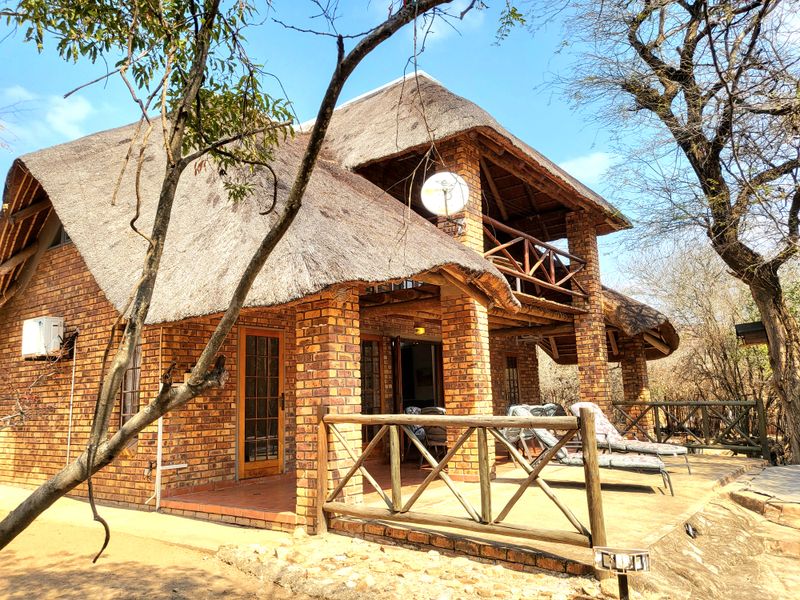A rustic double-story house featuring a traditional thatched roof, exposed brick exterior, and a spacious covered patio with a wooden balcony above.